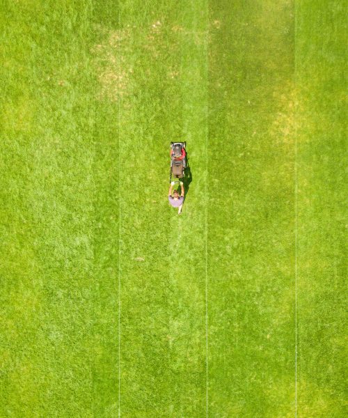 Aerial view of small figure of man worker trimming green grass with mowing mashine on football stadium field in summer. Aerial view of small figure of man worker trimming green grass with mowing mashine on football stadium field in summer.